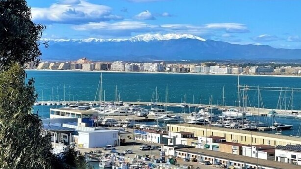 Una marina amb vaixells amarrats, un paisatge urbà costaner amb edificis, i muntanyes nevades al fons.