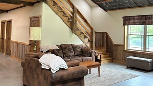 A spacious living area with a wooden staircase, a brown leather sofa, and a large window providing natural light. - Nature's Retreat: Ultimate ATV & Fishing Getaway minutes from Cherry Springs (Galeton)