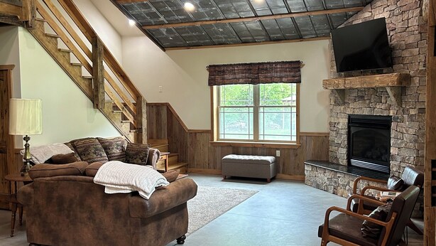 A spacious living room with a stone fireplace, a comfortable sofa, a wooden staircase, and a large window. - Nature's Retreat: Ultimate ATV & Fishing Getaway minutes from Cherry Springs (Galeton)