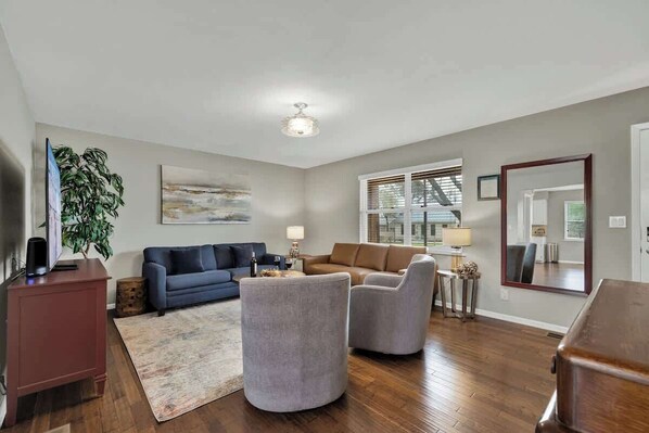 A spacious living room with a blue sofa, a brown leather sofa, and two grey armchairs. There is a wooden coffee table, a TV on a stand, and a large mirror on the wall.