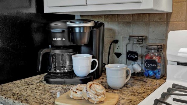 Eine Küche mit einer Keurig-Kaffeemaschine, zwei weißen Tassen, einem Schneidebrett mit Zimtschnecken und Gläsern mit den Aufschriften 'Sahne' und 'Kaffee'.