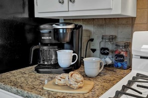 A kitchen with a Keurig coffee maker, two white mugs, a cutting board with cinnamon rolls, and jars labeled "Cream" and "Coffee".