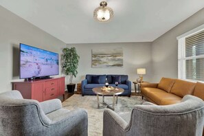 Ein Wohnzimmer mit Flachbildfernseher, einer Couch, zwei Sesseln, einem Couchtisch, einer Pflanze und Kunstwerken an den Wänden.