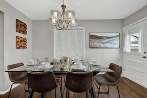 A dining area with a glass table, four chairs, a chandelier, and a painting on the wall.