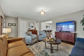 A living room with a sofa, armchairs, a coffee table, a TV, and a dining area in the background.