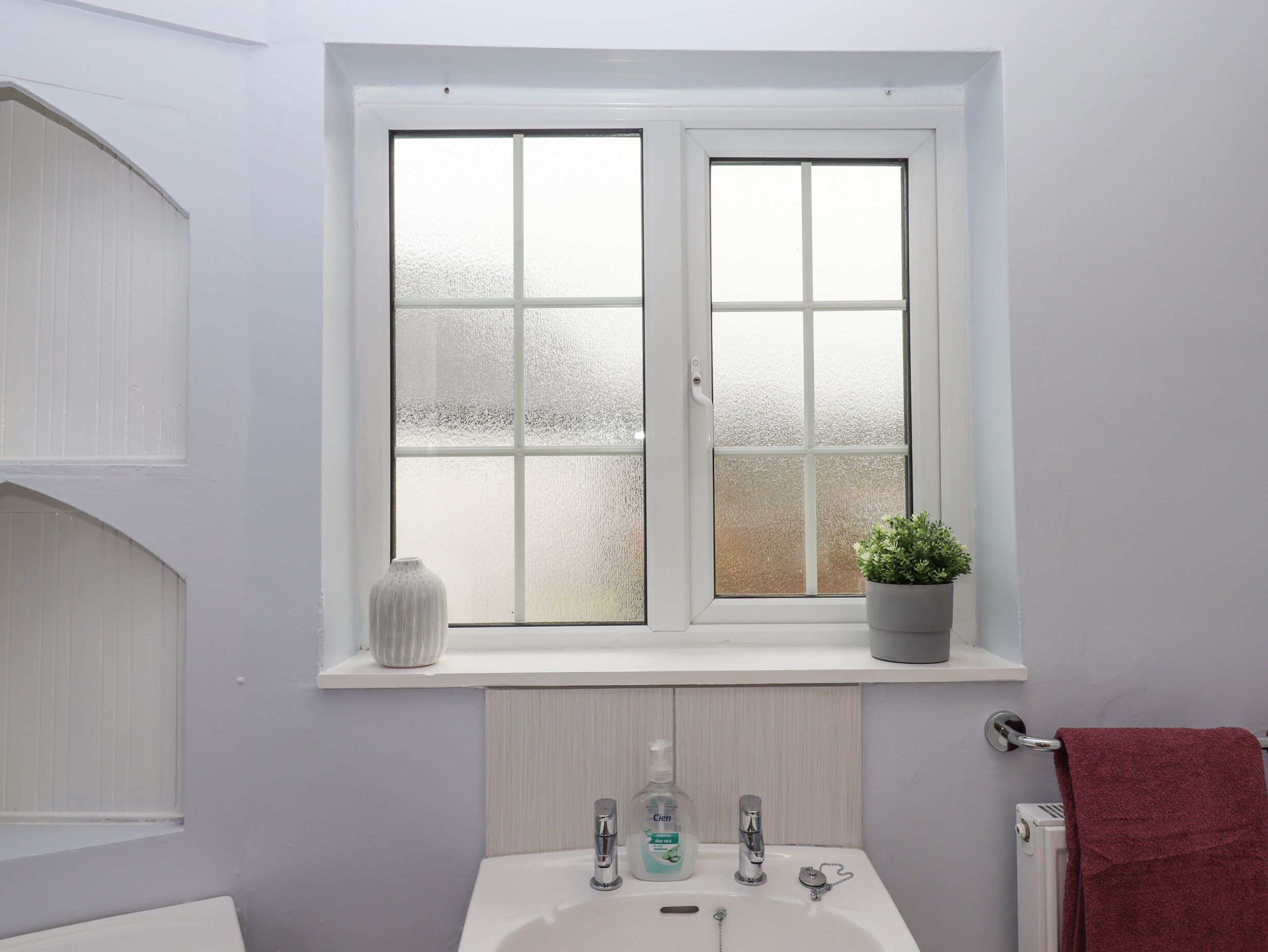 A bathroom with a white sink, a window with frosted glass, a potted plant, and a red towel.