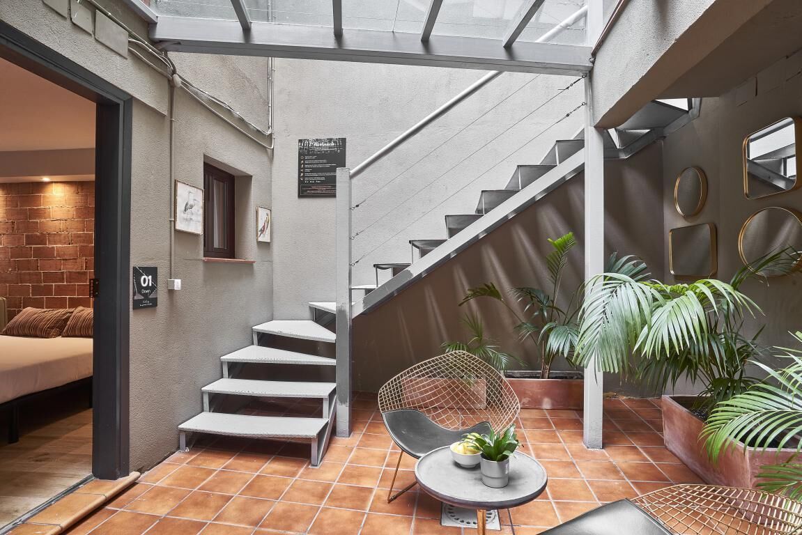 Un interior moderno con una escalera, una cama, una zona de comedor con mesa y sillas, y plantas en macetas.