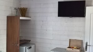 Une petite pièce aux dimensions réduites, avec des murs en bois peints en blanc, un téléviseur à écran plat fixé au mur, une armoire en bois, un petit réfrigérateur et une boîte remplie d’objets posée sur le sol.