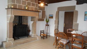 A room with a stone fireplace, wooden chairs, and a dining table. - Gîte 5 Personnes (Le Vast)