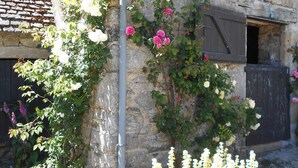 A stone building with a rustic appearance, featuring a metal wheel and flowering plants. - Gîte 5 Personnes (Le Vast)