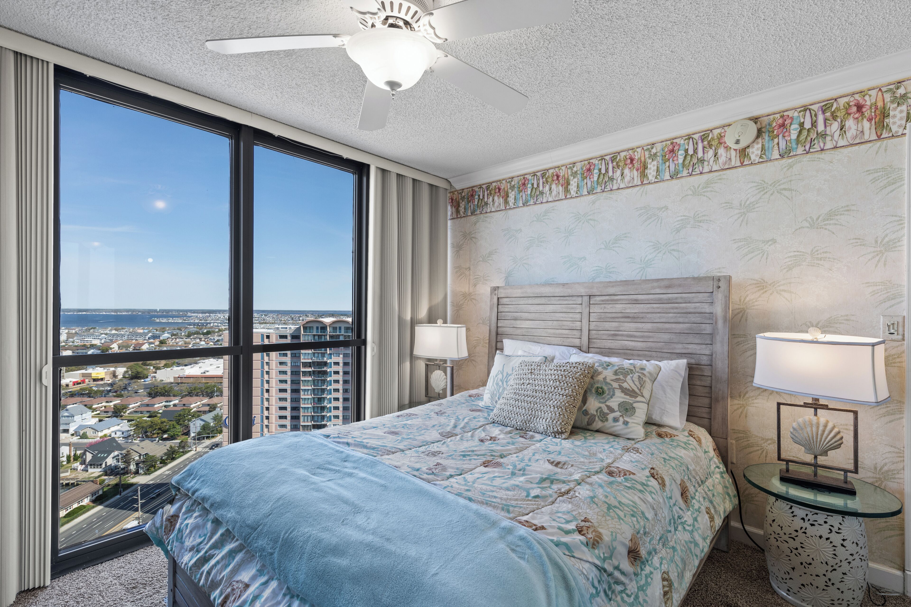 A hotel room with a large window, a bed with a floral bedspread, a ceiling fan, and a nightstand with a lamp.