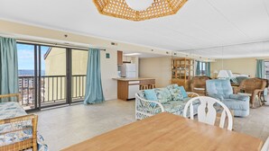 A spacious living area with a balcony, wicker furniture, a wooden dining table, and a kitchen area. - Summer Beach 801 by Seaside Vacations & Sales (Ocean City)