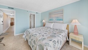 A bedroom with a bed, two bedside tables with lamps, a painting on the wall, and a hallway leading to another room. - Summer Beach 801 by Seaside Vacations & Sales (Ocean City)