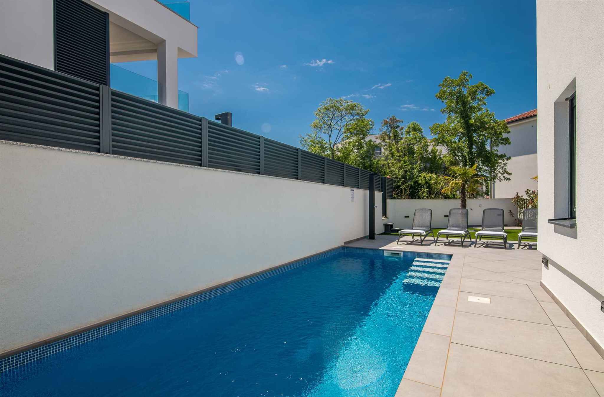 A modern pool area with a clear blue pool, a white wall, and a row of chairs.