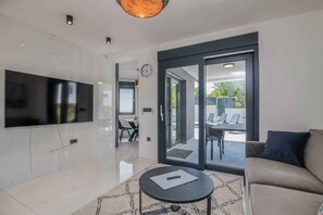 A modern living room with a flat-screen TV, a sofa, a coffee table, and a sliding glass door leading to an outdoor seating area. - Holiday apartment with pool and air conditioning (Njivice)