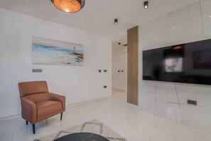 A modern living room with a leather armchair, a flat-screen TV mounted on the wall, and a painting of a seascape.