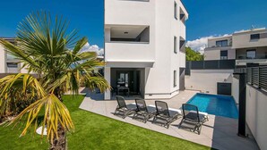 Un edificio moderno de varios pisos con piscina, mobiliario exterior y una palmera.