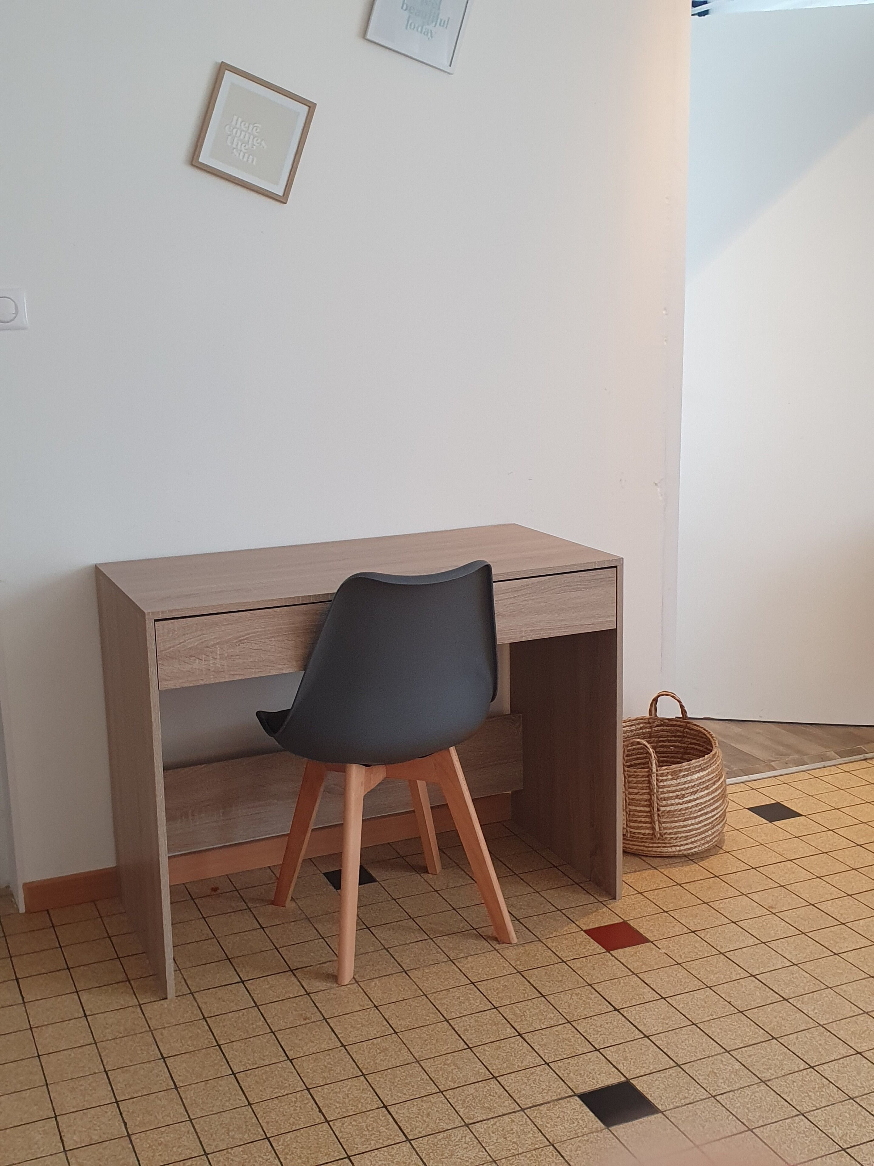 Une petite pièce, bien rangée, avec un bureau en bois, une chaise noire, un panier en osier et un sol carrelé.