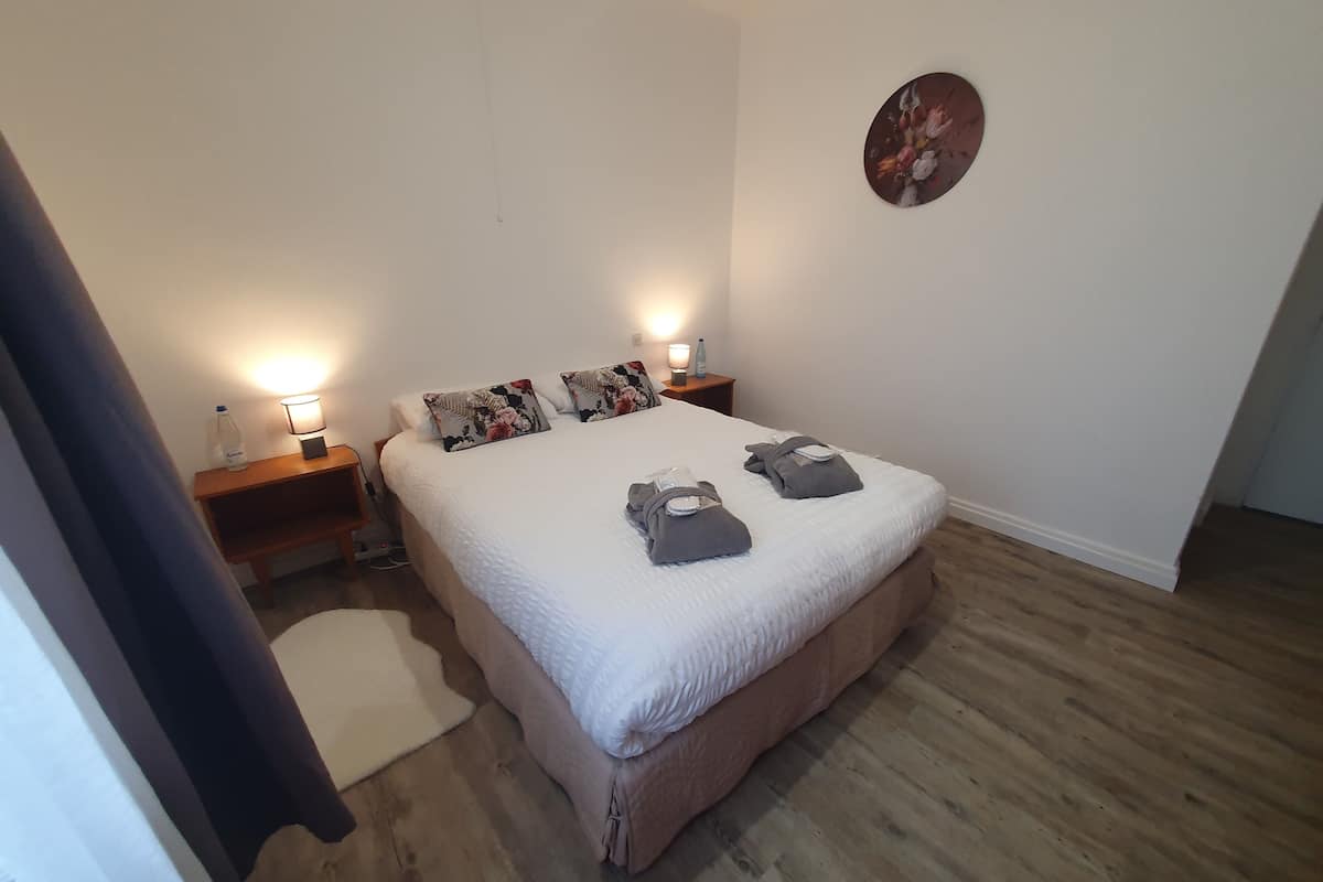 A neatly arranged bedroom with a large bed, two bedside tables with lamps, a wall clock, and a wooden floor.