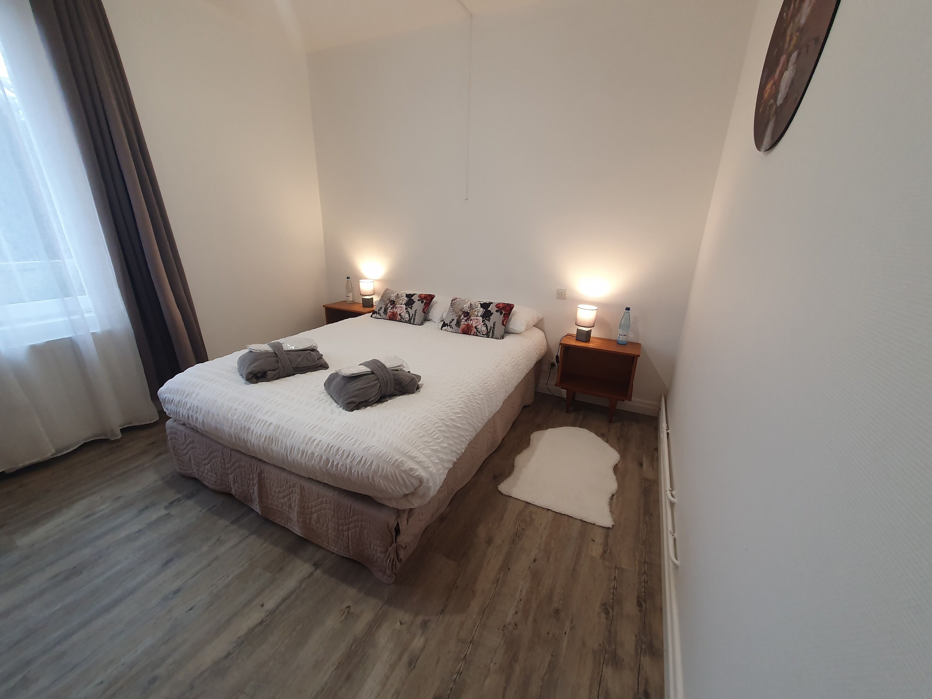 En pænt redt seng med to puder, et sengebord med en lampe, og en lille måtte på et trægulv.