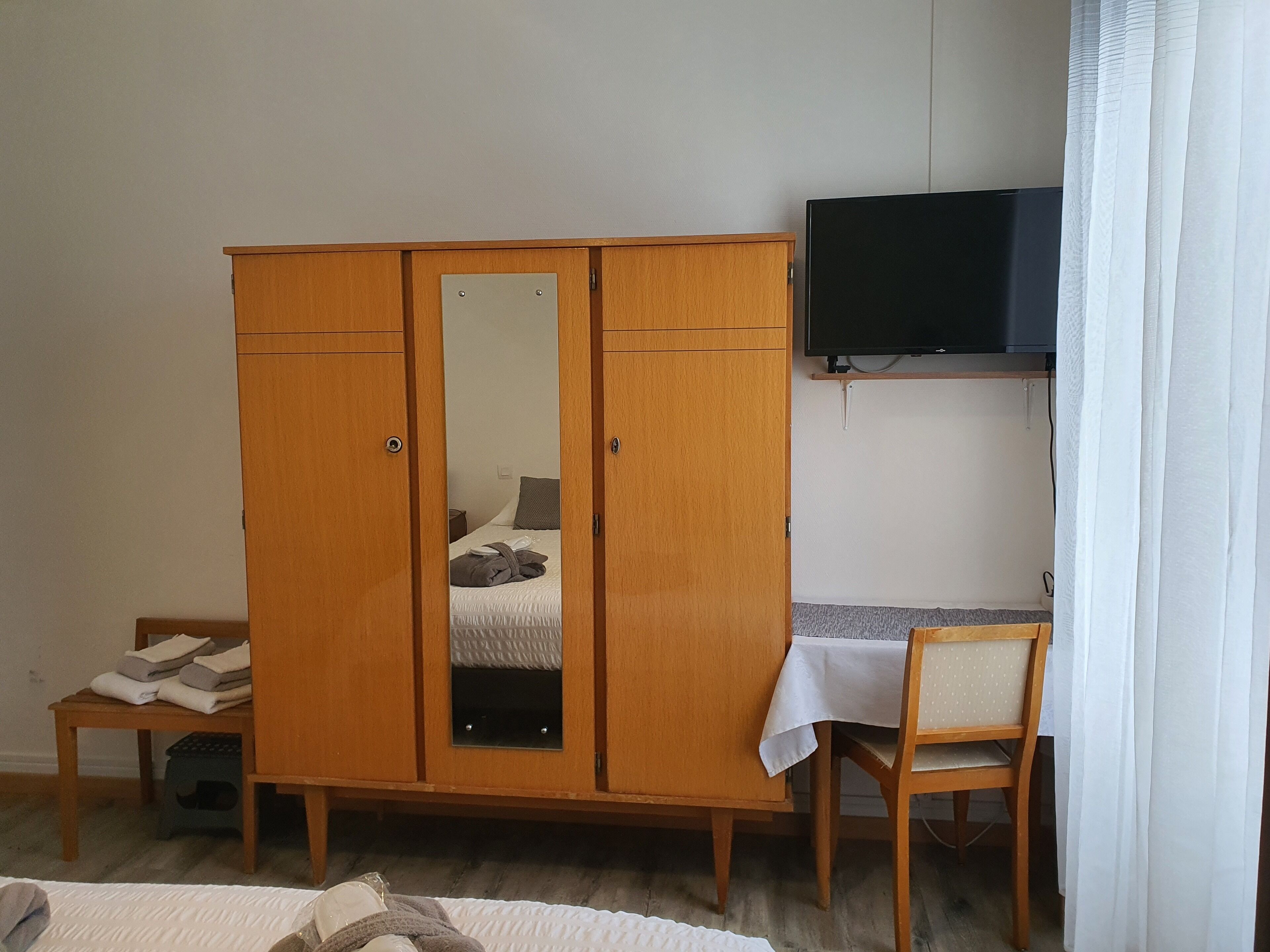 Een slaapkamer met een houten garderobe, een bed met een witte dekbedovertrek, een klein tafeltje met een witte tafelkleed, een houten stoel en een televisie aan de muur.