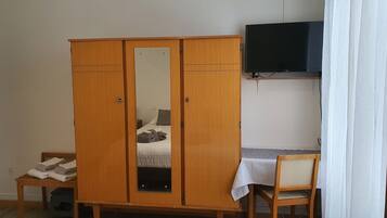 Een slaapkamer met een houten garderobe, een bed met een witte dekbedovertrek, een klein tafeltje met een witte tafelkleed, een houten stoel en een televisie aan de muur.