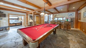 A pool table in a room with stone flooring, wooden beams, and a bar area with stools.