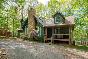 A cabin with a stone chimney, a porch, and a gravel driveway surrounded by trees.