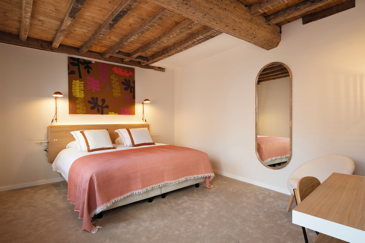 A bedroom with a wooden ceiling, a large bed with white and pink bedding, a wall-mounted mirror, and a small table.
