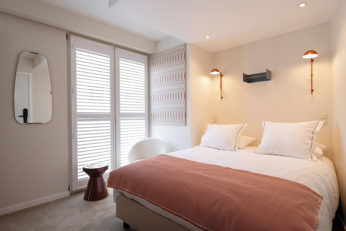 A bedroom with a bed, a chair, a mirror, and a wall-mounted light fixture.