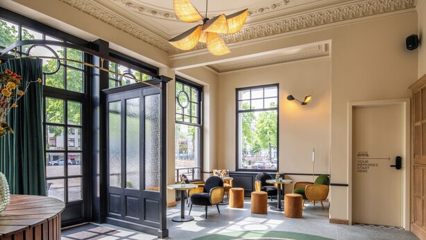 A hotel lobby with a large black door, multiple seating areas, and a ceiling fan. - Made in Catherine (Brussels)