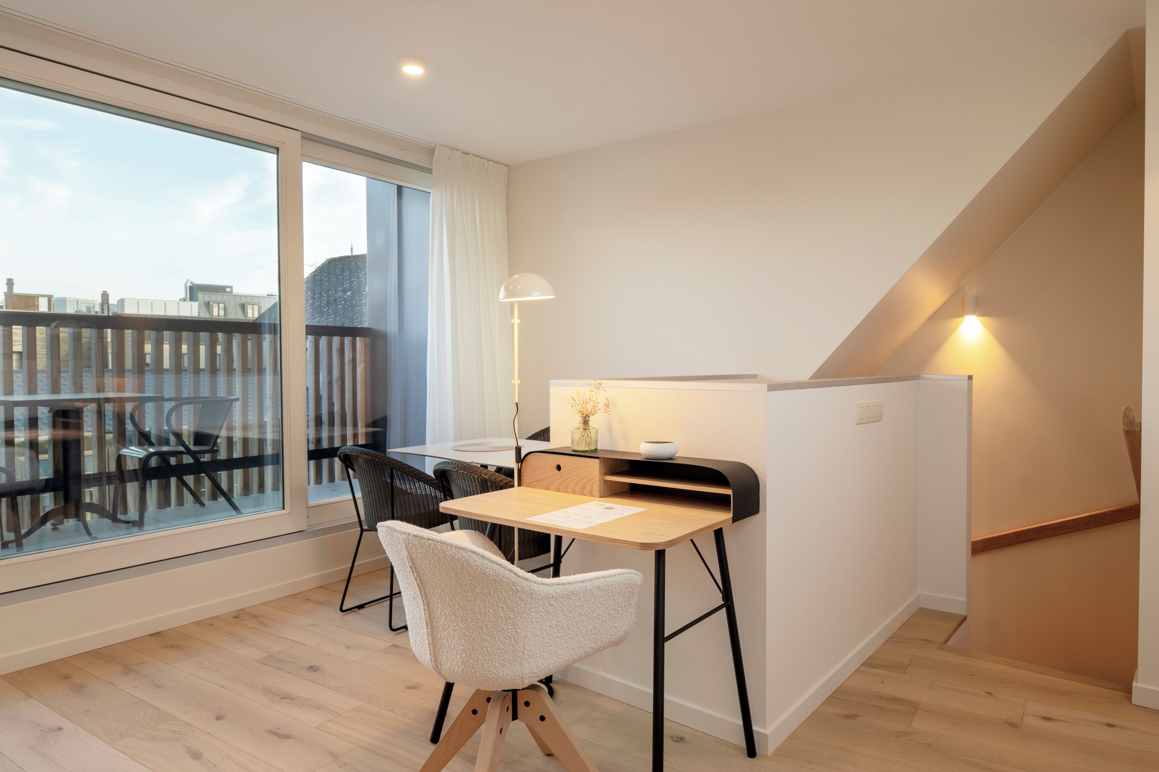 Ein modernes Zimmer mit Schreibtisch, zwei Stühlen, einer Lampe und Blick auf die Stadt durch große Fenster.