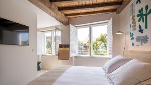 A bedroom with a bed, a window with shutters, a wall-mounted TV, and a wooden ceiling beam. - Made in Catherine (Brussels)