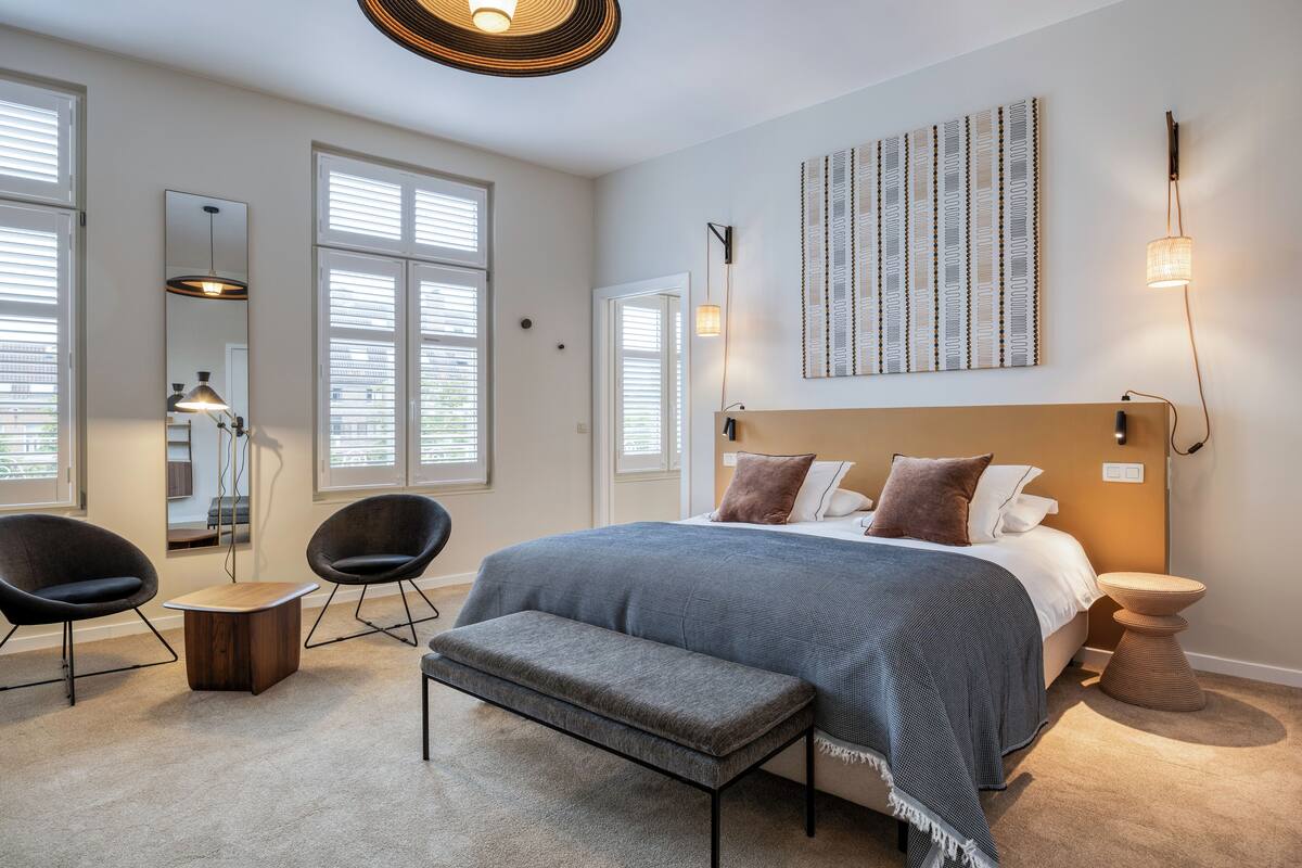 A modern bedroom with a large bed, two chairs, a bench, a bedside table, and a wall-mounted light fixture.