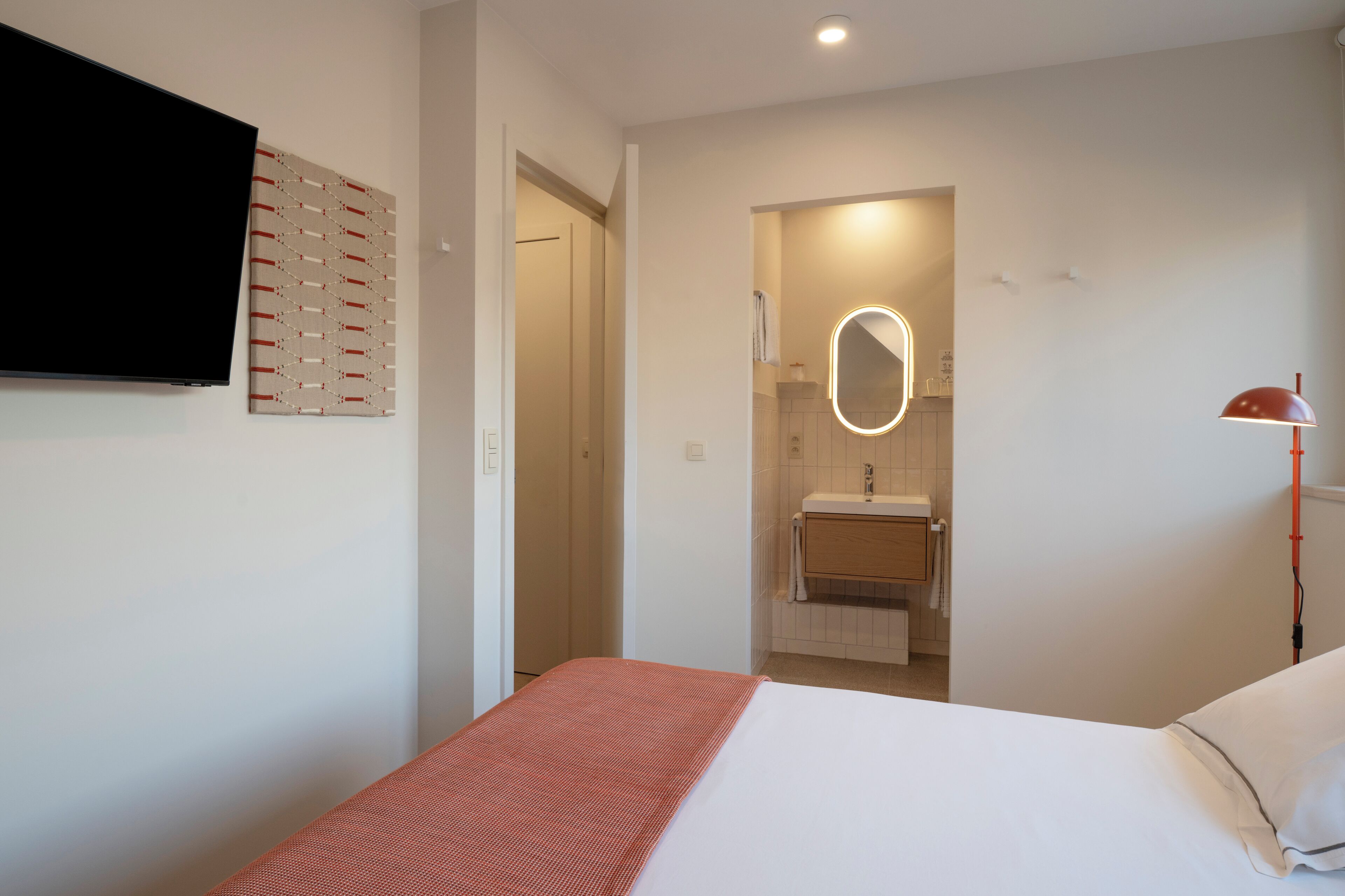 A bedroom with a bed, a TV mounted on the wall, a bathroom with a mirror, and a red lamp.