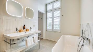 A modern bathroom with a white sink, vanity, and bathtub. There are two illuminated mirrors above the sink and a window with blinds to the right.