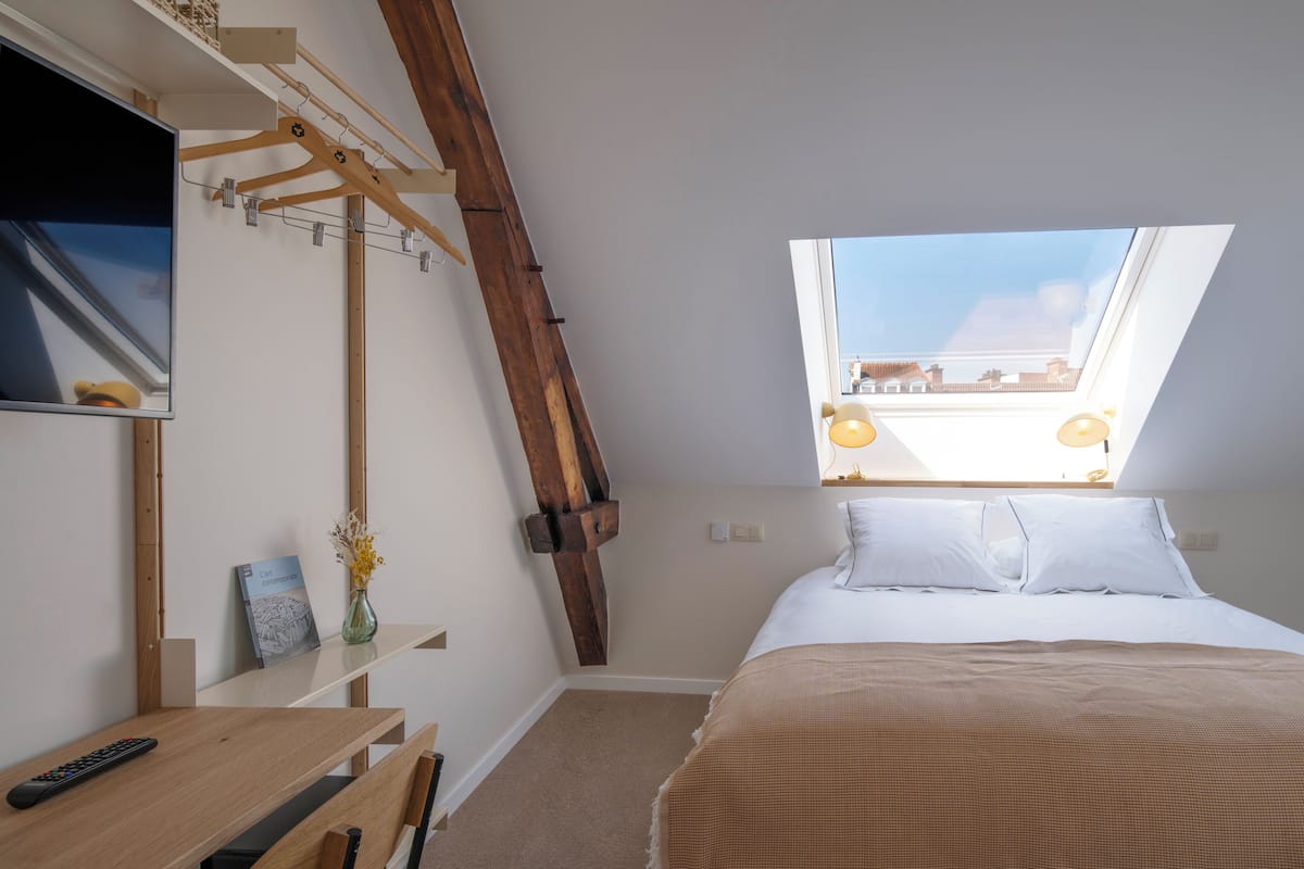 A bedroom with a slanted ceiling, a bed with white bedding, a wooden desk, a remote control, a vase with flowers, and a skylight.
