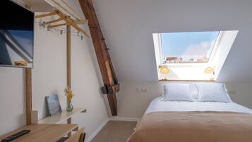 Un dormitorio con techo inclinado, cama con ropa de cama blanca, un escritorio de madera, un control remoto, un jarrón con flores y un tragaluz.