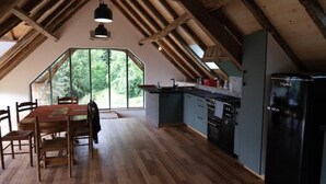 Un coin repas avec une table et des chaises en bois, une cuisine équipée d’un réfrigérateur et d’une cuisinière, et une grande fenêtre donnant sur la forêt.