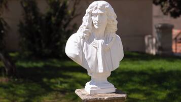 A white bust sculpture on a stone pedestal in a garden setting.