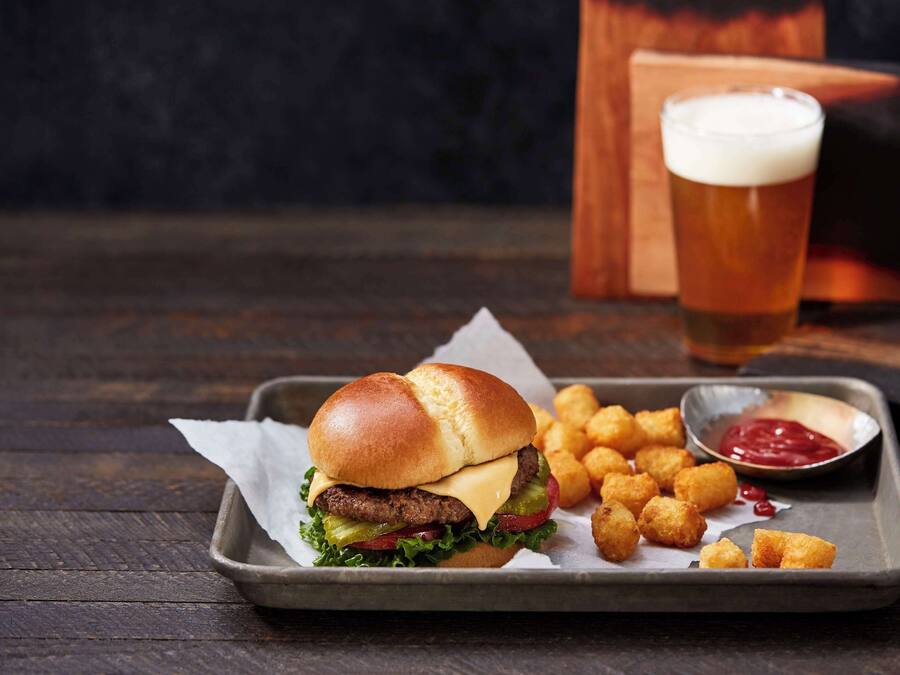 A burger with fries and ketchup on a tray, a glass of beer, and a dark background.