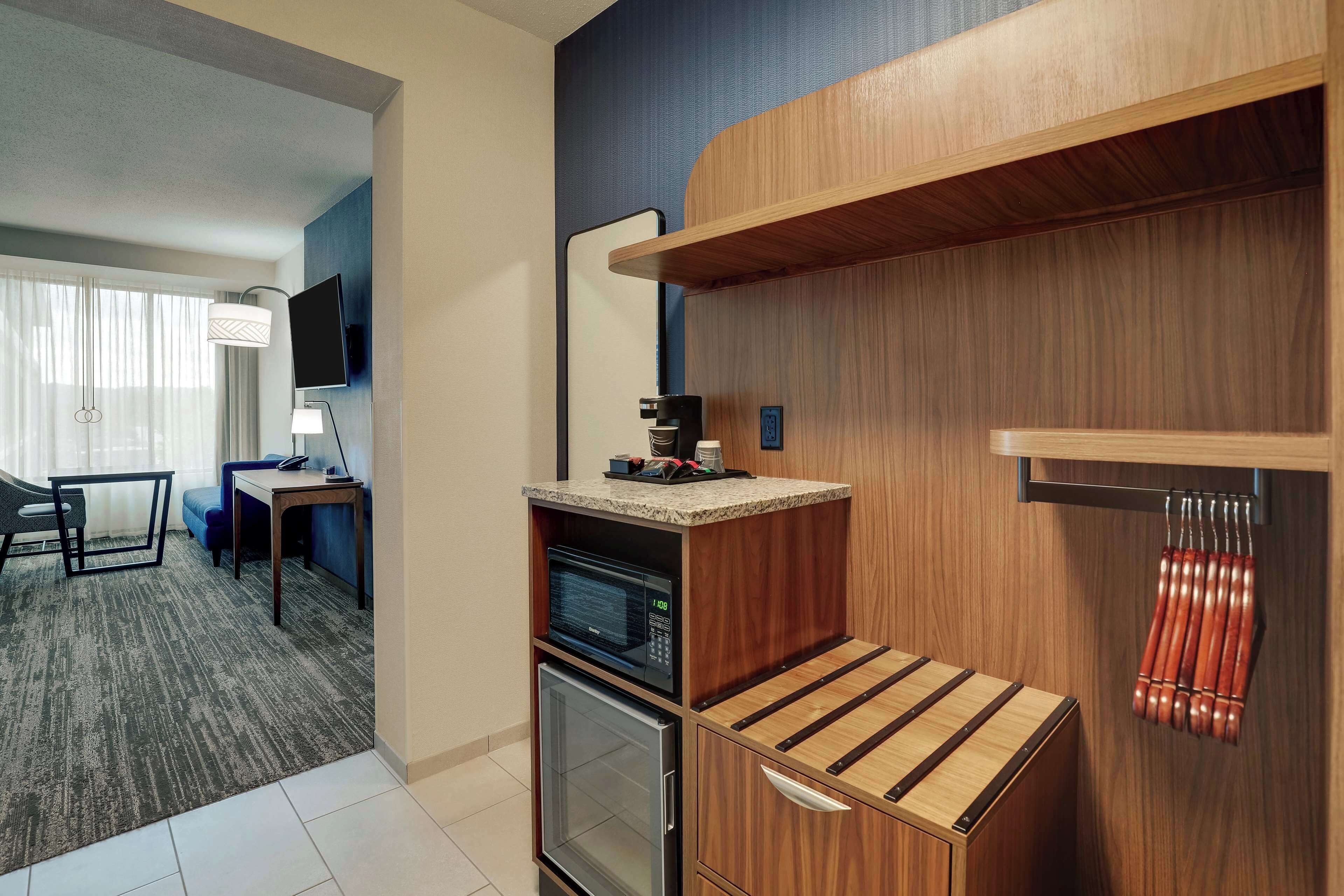A compact hotel room with a kitchenette, a small refrigerator, a microwave, a coffee maker, and a TV mounted on the wall.