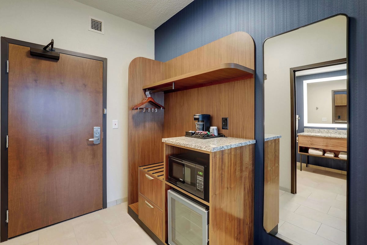 Ein kleines Hotelzimmer mit Holztür, einer Mikrowelle, einem kleinen Kühlschrank, einem Holzboden und einer Kaffeemaschine sowie einem Spiegel.