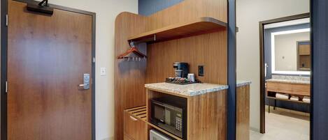 A compact hotel room with a wooden door, a microwave, a small refrigerator, a wooden shelf with a coffee maker, and a mirror.
