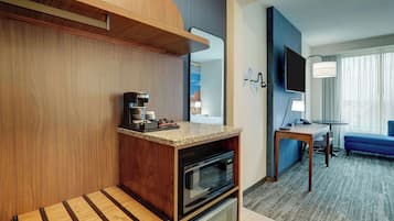 A compact hotel room with a kitchenette, a small dining area, a TV, and a window with curtains.