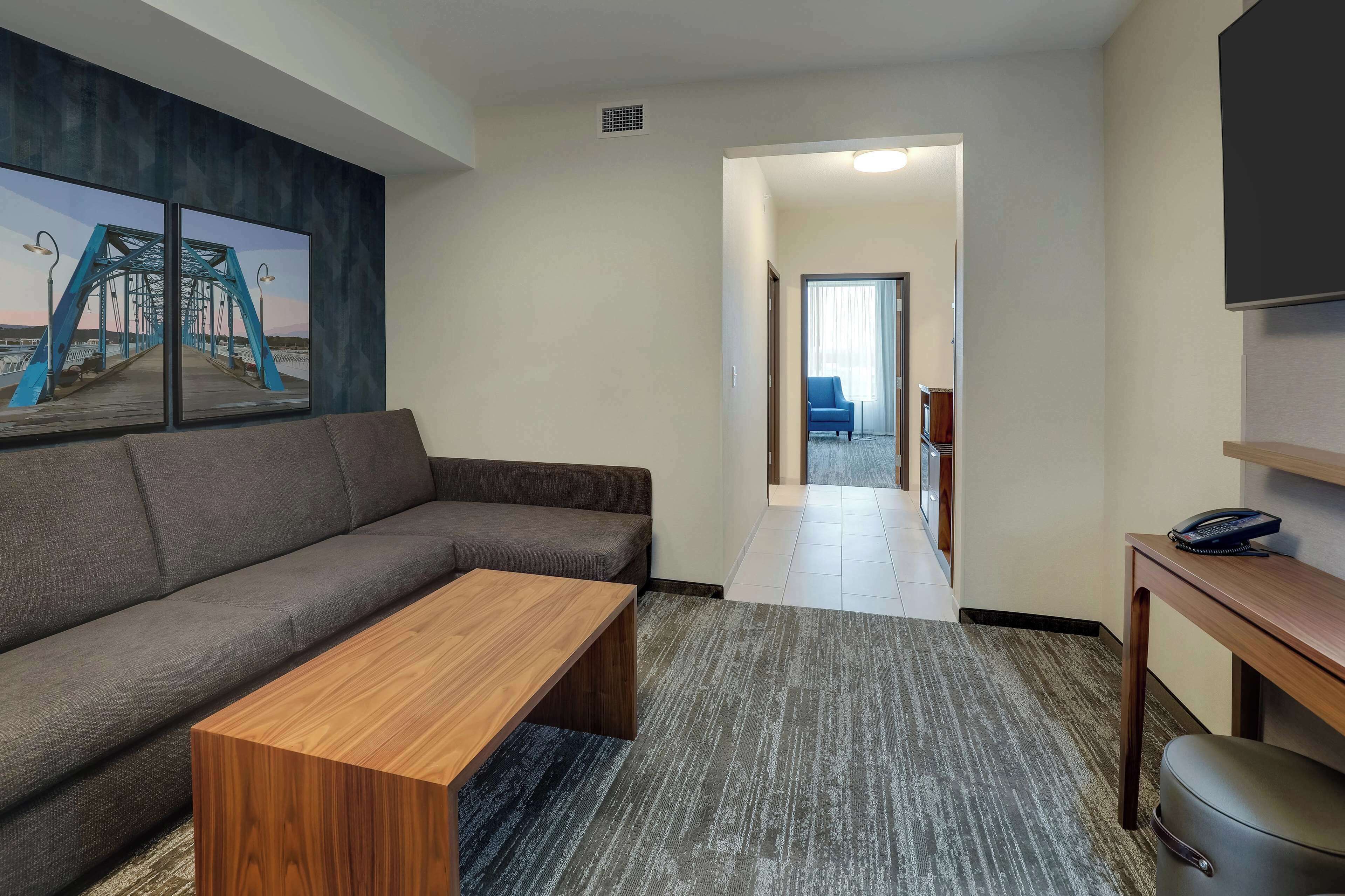 A modern hotel room with a gray sofa, a wooden coffee table, and a large painting of a bridge.