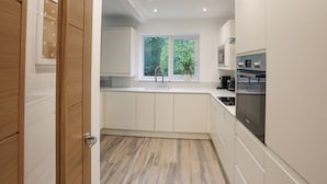 A modern kitchen with white cabinets, a stainless steel oven, and a window with a view of greenery. - 15 Maes Awel (Pwllheli)