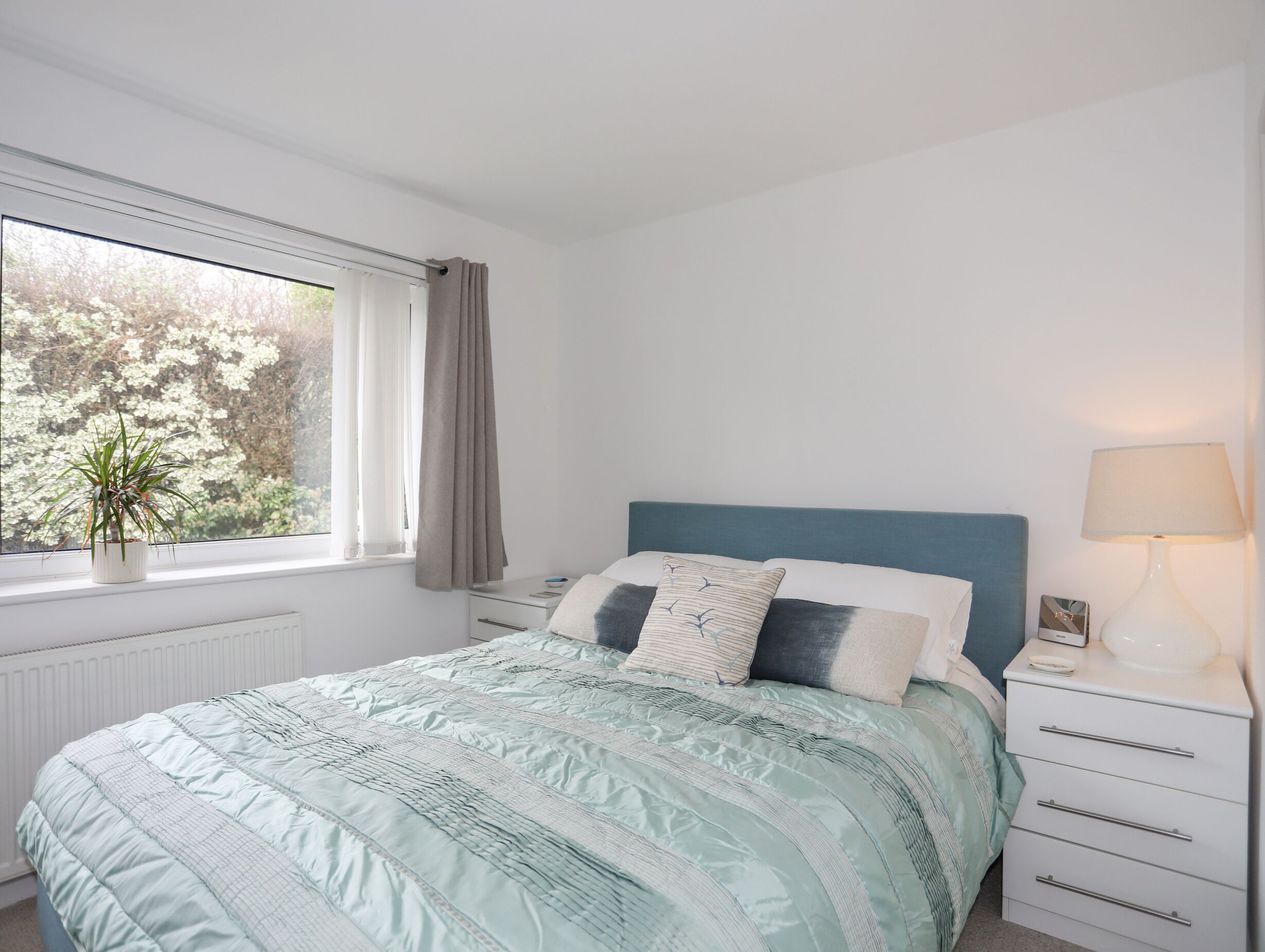 A bedroom with a bed, a nightstand, a lamp, a window with a view of trees, and a potted plant.