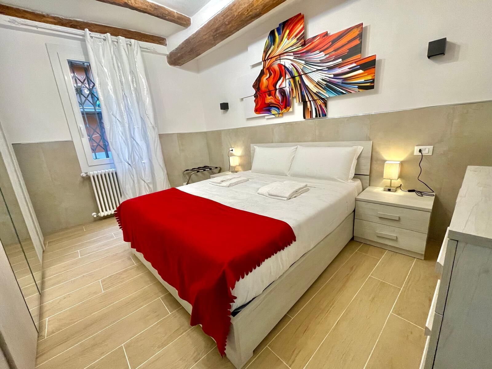 Une chambre moderne avec un grand lit, un plaid rouge, une tête de lit, des lampes de chevet et une fenêtre avec des rideaux.