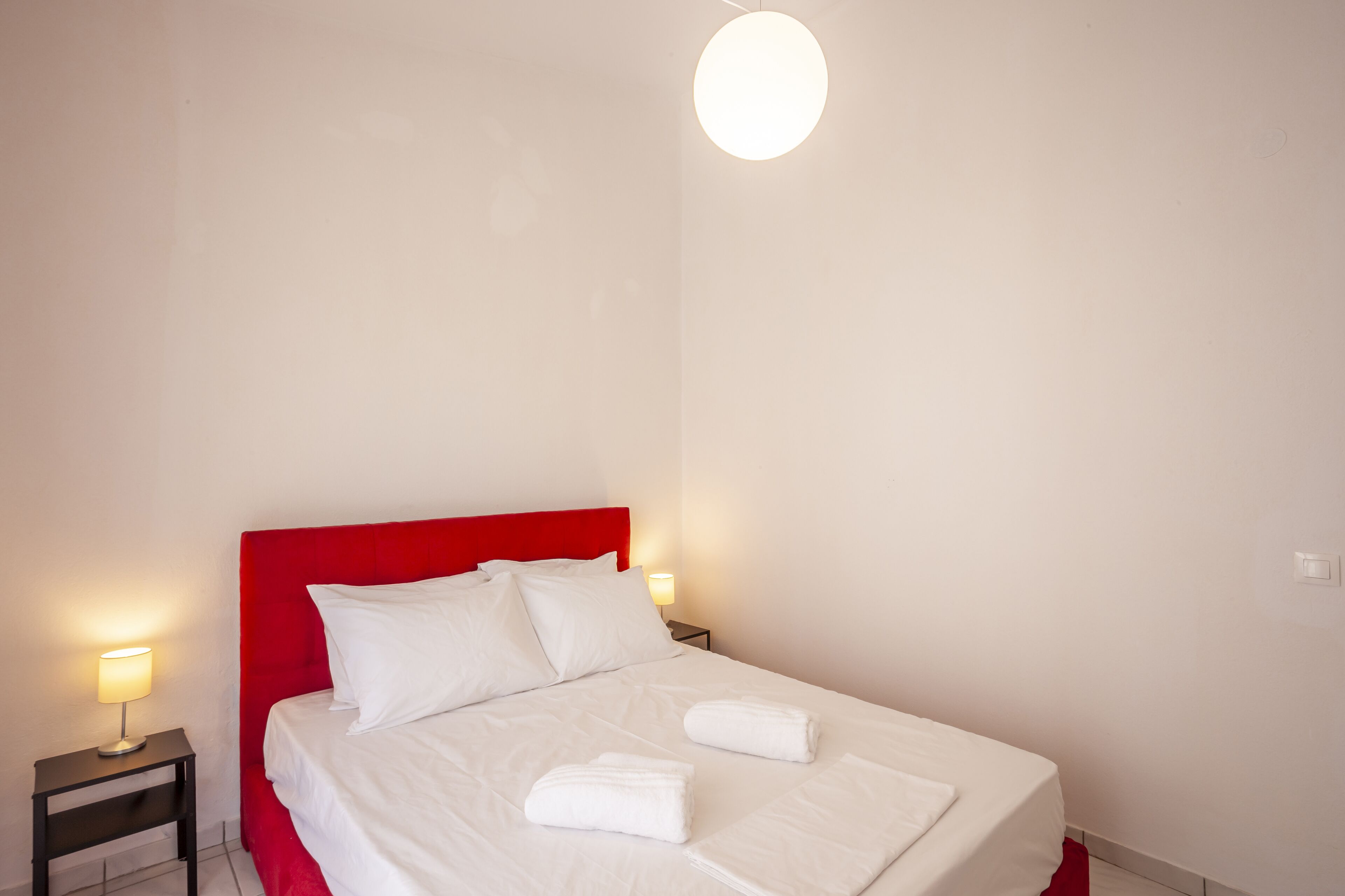A neatly made bed with white linens and two towels, a red headboard, and a bedside lamp on a nightstand.